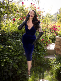 Micheline Pitt, posing with one hand on her face and the other pressed against her thigh, models the Baudelaire Wiggle Dress in Midnight Blue paired with elbow length black gloves.