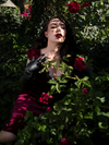Slightly obscured by a rose bush, Micheline Pitt models the  Baudelaire Wiggle Dress in Oxblood.