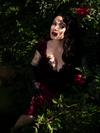 Micheline Pitt sitting in a shady garden setting surrounded by pink roses models the  Baudelaire Wiggle Dress in Oxblood.