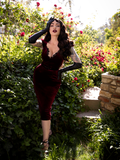Micheline Pitt, holding her right hand up to her temple, models an oxblood color gothic style dress from La Femme en Noir.