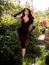 Micheline Pitt wearing the  Baudelaire Wiggle Dress in Oxblood while standing in a lush garden setting.