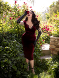 Micheline Pitt, holding a rose up to her eye that matches the color of her  Baudelaire Wiggle Dress in Oxblood.