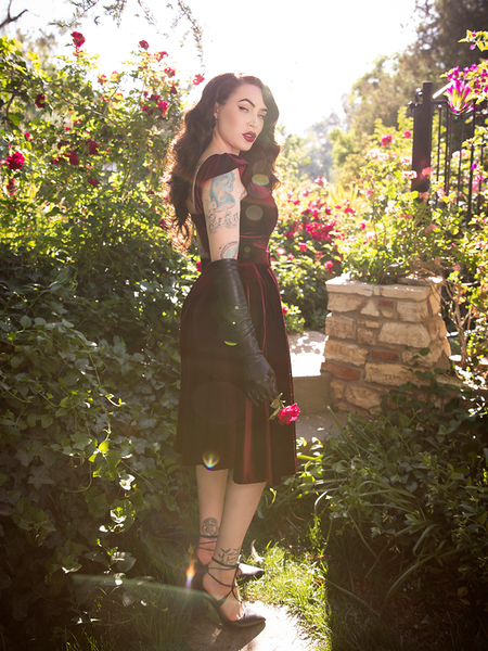 Turned to the side while holding a pink rose, Micheline Pitt stuns in the Baudelaire Swing Dress in Oxblood from gothic dress company La Femme en Noir.