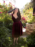 Holding her hand up to her temple, Micheline Pitt gazes into the camera while modeling the Baudelaire Swing Dress in Oxblood from gothic glamour clothing company La Femme en Noir.
