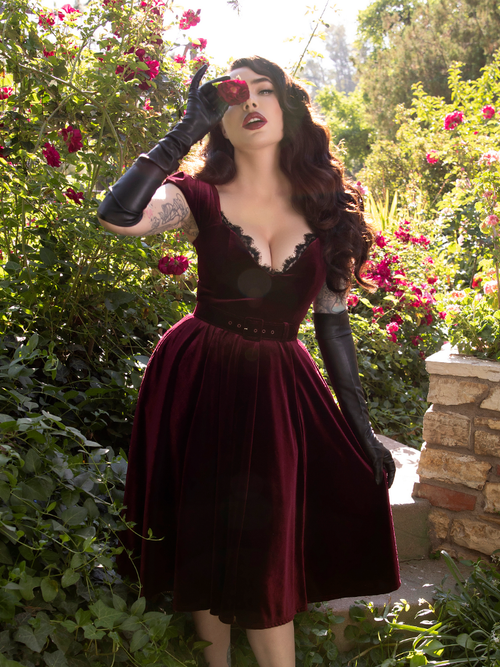 Micheline Pitt standing in a rose garden while wearing the Baudelaire Swing Dress in Oxblood from gothic retro clothing brand La Femme en Noir.