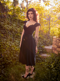 Stephanie stands in a sunny garden while wearing the Baudelaire Swing Dress in Black.