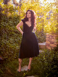 Stephanie posing while standing on a stone pathway in the middle of a lush green garden while modeling an all new goth style dress from La Femme en Noir - Baudelaire Swing Dress in Black.