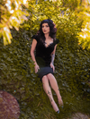 Stephanie sitting amongst a wall of thick, green ivy wears the Baudelaire Wiggle Dress in Black from goth retro clothing company La Femme en Noir.