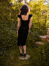 The back of the Baudelaire Wiggle Dress in Black as worn by Stephanie Joens in a lush, green garden setting.