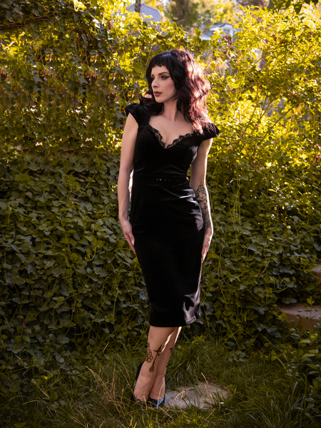 Standing in a green garden being illuminated by the sun, Stephanie wears an all new goth style dress from La Femme en Noir called the Baudelaire Wiggle Dress in Black.