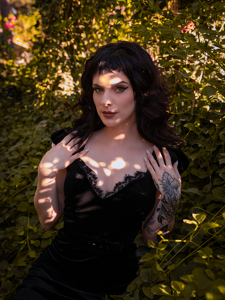 Stephanie posing seductively in the Baudelaire Wiggle Dress in Black in an outdoor area lush garden setting.