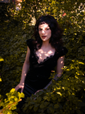 Stephanie wears the Baudelaire Wiggle Dress in Black while sun breaks through the rose bushes surrounding her.
