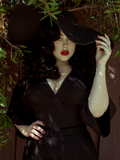 Rachel stands under a tree with her hand on a black wide brim hat while modeling the Bauhaus top in black from La Femme En Noir.