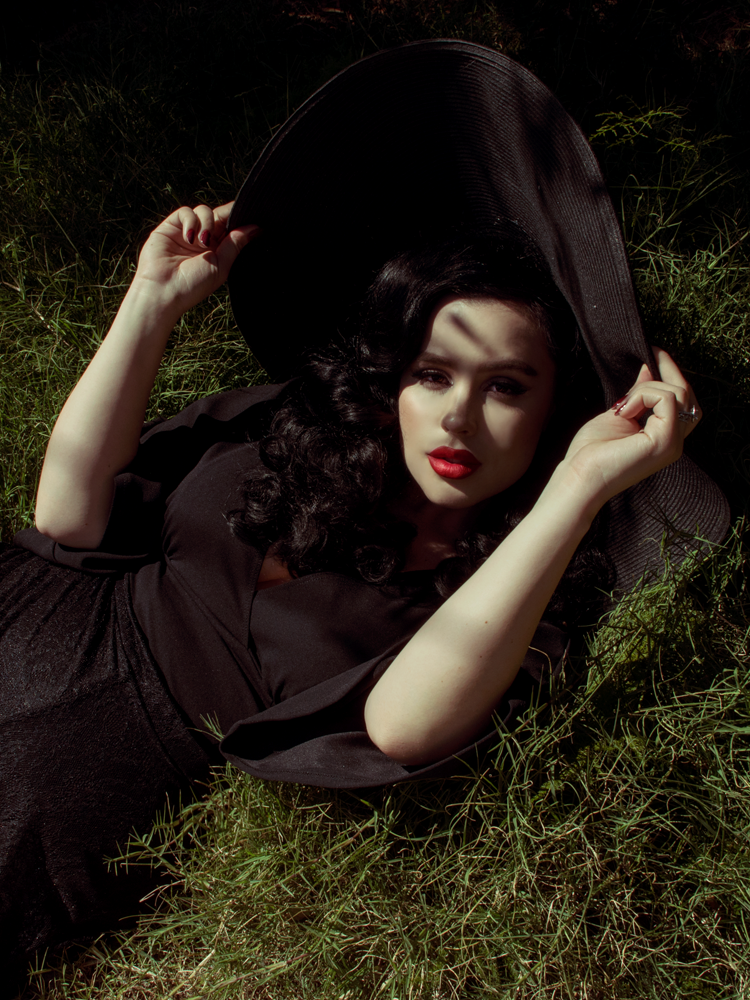 Rachel, relaxing in grass with her hands on a black wide brim hat, models the Bauhaus top in black from La Femme En Noir.
