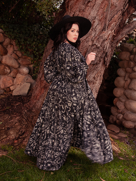 The mesmerizing brunette model effortlessly rocks the Belladonna Maxi Dress in Cottage Witch Toile Print from the gothic clothing brand La Femme en Noir in a garden setting.