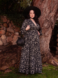 Surrounded by nature's beauty, the lovely brunette model exhibits the Belladonna Maxi Dress in Cottage Witch Toile Print from the gothic clothing brand La Femme en Noir.
