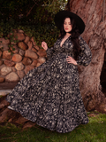 Amidst a garden scene, the stunning brunette model flaunts the Belladonna Maxi Dress in Cottage Witch Toile Print from the gothic clothing brand La Femme en Noir.