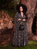 Embracing the enchanting atmosphere of a garden, the brunette model showcases the Belladonna Maxi Dress in Cottage Witch Toile Print from the gothic clothing brand La Femme en Noir.