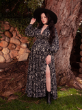 Posing in a garden setting, the gorgeous brunette model showcases the Belladonna Maxi Dress in Cottage Witch Toile Print from the gothic clothing brand La Femme en Noir.