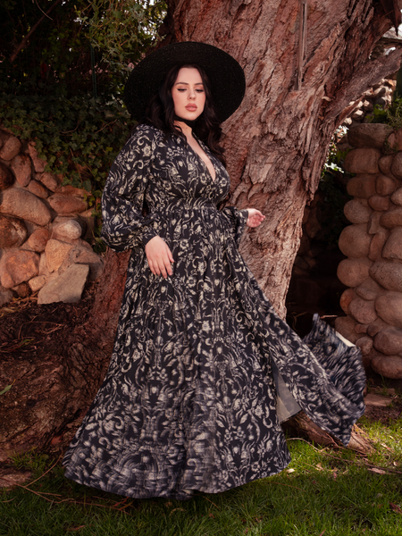 Captivating in a garden backdrop, the brunette model proudly presents the Belladonna Maxi Dress in Cottage Witch Toile Print from the gothic clothing brand La Femme en Noir.