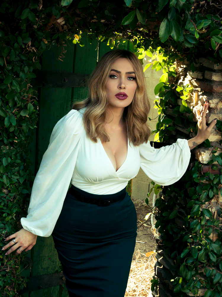 Micheline Pitt enters a lush garden while modeling the Bishop blouse in ivory by La Femme En Noir.