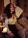 Micheline Pitt sits in a gothic bedroom while modeling the Bishop blouse in ivory by La Femme En Noir.