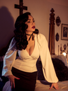 Micheline Pitt leans against a bed in a gothic bedroom while modeling the Bishop blouse in ivory by La Femme En Noir.