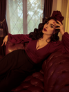 Micheline Pitt lounges on a tufted leather couch while modeling the Bishop blouse in oxblood by La Femme En Noir.