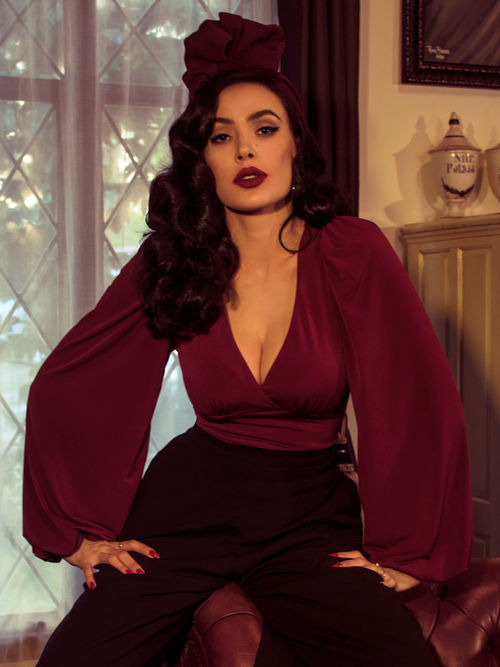 Micheline Pitt sits on a tufted leather couch while modeling the Bishop blouse in oxblood by La Femme En Noir.