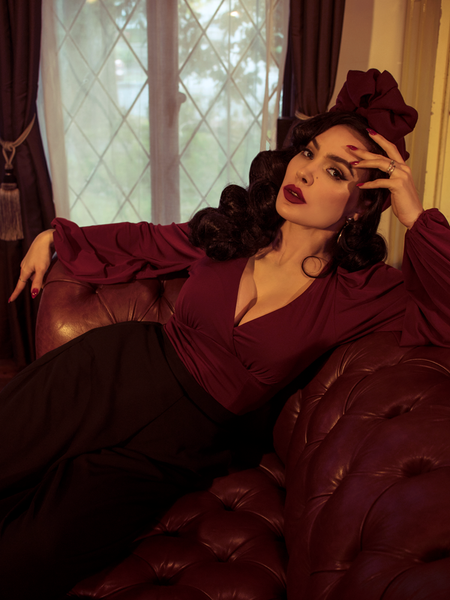 Micheline Pitt lounges on a tufted leather couch while modeling the Bishop blouse in oxblood by La Femme En Noir.