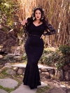 Rachel Sedory standing with her hand on her hip and one gently pulling on her hair while wearing the Black Marilyn Lace Gown in Black.