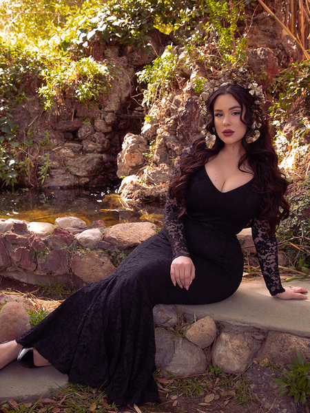 Rachel Sedory sat on a rock wall in front of a pond wearing the Black Marilyn Lace Gown in Black.