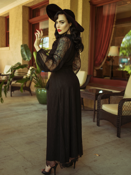 Micheline Pitt with her body facing away from the camera, subtly turns her head back while wearing the Black Widow Wrap Gown in Black Lace. 
