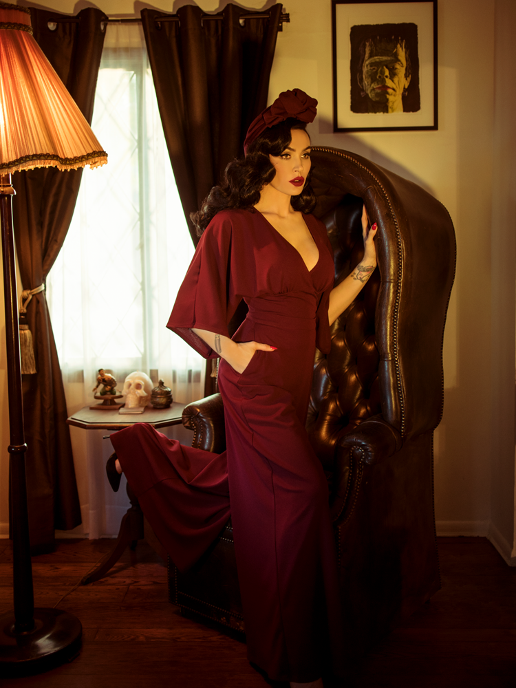 Micheline Pitt stands against a gothic style leather chair while modeling the Black Widow palazzo pants in oxblood by La Femme En Noir.
