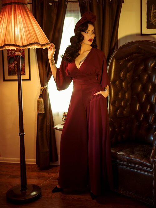 Micheline Pitt standing next to a gothic style leather chair while modeling the Black Widow palazzo pants in oxblood by La Femme En Noir.