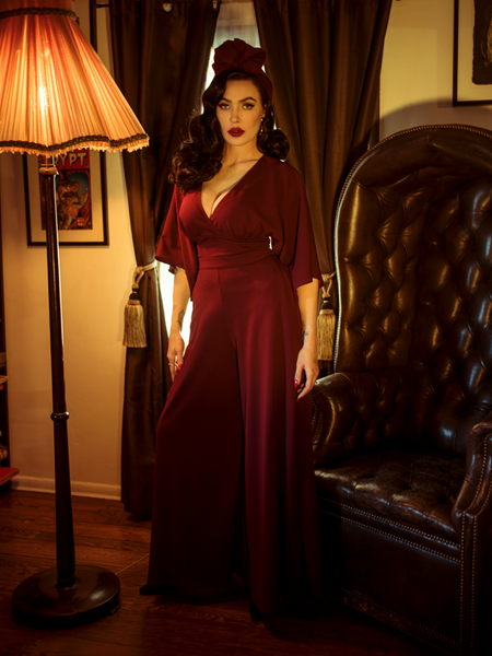 Micheline Pitt standing next to a gothic style leather chair while modeling the Black Widow palazzo pants in oxblood by La Femme En Noir.