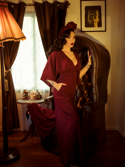 Micheline Pitt stands against a gothic style leather chair while modeling the Black Widow palazzo pants in oxblood by La Femme En Noir.