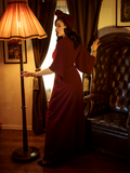 A back shot of Micheline Pitt standing near a gothic style leather chair while modeling the Black Widow palazzo pants in oxblood by La Femme En Noir.