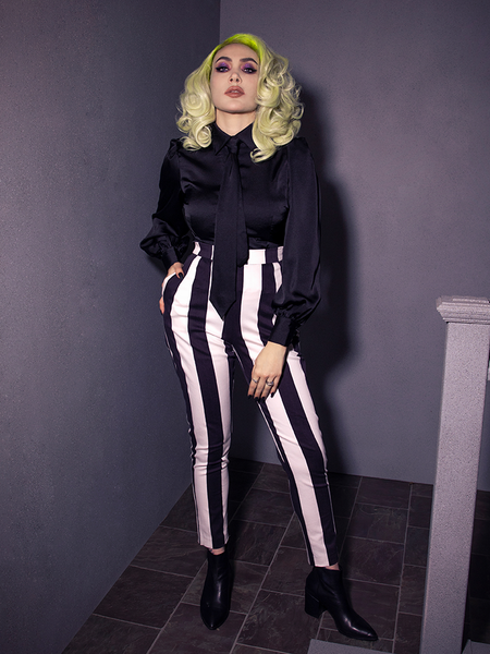 Full length shot of Micheline Pitt standing and posing in the Bowie Blouse w/Matching Tie in Black Charmeuse being worn tucked into black and white striped pants.