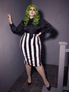 Full length image of female with green hair wearing a long sleeve black shiny top tucked into a black and white striped skirt. The outfit is finished off with black flats.