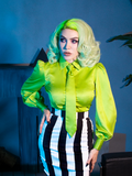Micheline Pitt posing in front of a spooky scene while wearing the Bowie Blouse w/Matching Tie in Lime Charmeuse with black and white striped skirt.