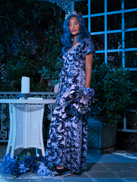 Vanessa holding a bouquet of blue roses standing in Tim Burton's CORPSE BRIDE™ Butterfly Maxi Dress in Dusk Blue from goth glamour clothing company La Femme en Noir.