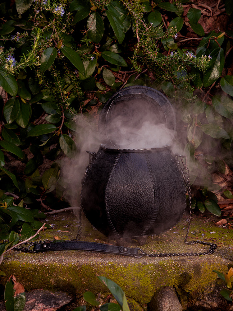 The Cottage Witch Cauldron Crossbody Bag, created by La Femme en Noir's gothic glamour clothing brand, looks enchanting in the garden setting.