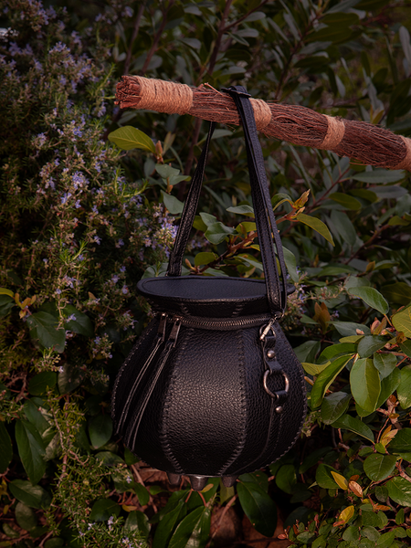 La Femme en Noir's Cottage Witch Cauldron Crossbody Bag, captured in a beautiful garden setting, is the epitome of gothic glamour.