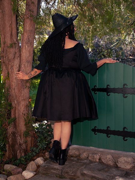 Within the depths of the shady woodland, a stunning fair-skinned brunette figure embodies the essence of gothic style, showcasing the bewitching Cottage Corset Pinafore in Black, a must-have accent piece for your gothic dress wardrobe.