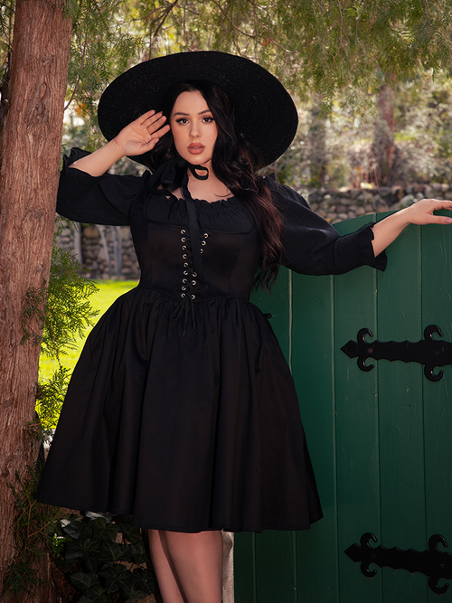 In the depths of the shadowy forest, a fair-skinned brunette model strikes a pose, showcasing the allure of the Cottage Corset Pinafore in Black, a must-have accent piece for any gothic dress connoisseur.