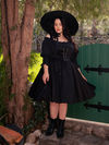 Lost in the mysterious woodland, a fair-skinned brunette muse captures the essence of gothic fashion, wearing the enchanting Cottage Corset Pinafore in Black, a versatile accent piece for your gothic dress ensemble.