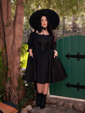 Amidst the haunting ambiance of the forest, a captivating fair-skinned brunette model exudes grace, adorned in the striking Cottage Corset Pinafore in Black, an exquisite complement to your gothic dress collection.