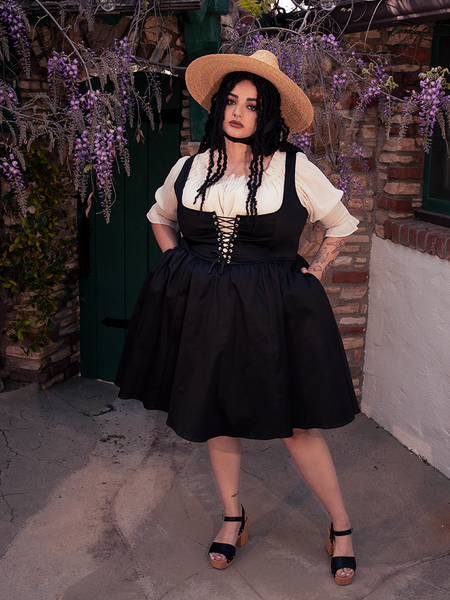 In the mystical embrace of the forest, a fair-skinned brunette beauty emanates gothic sophistication, adorned in the enchanting Cottage Corset Pinafore in Black, a versatile accent piece that enhances your gothic dress ensemble.