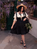 Embraced by the eerie ambiance of the forest, a captivating fair-skinned brunette model exudes a sense of gothic allure, donning the exquisite Cottage Corset Pinafore in Black, an essential addition to your gothic dress ensemble.